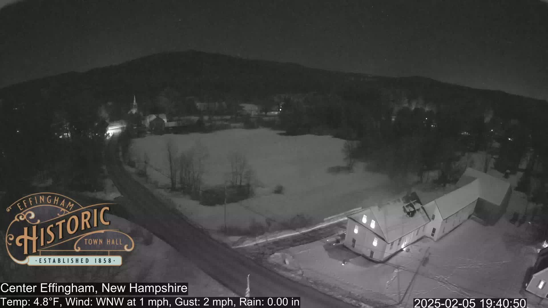 A nighttime, snowy scene shows a small town with a few buildings and roads, under a dark sky with minimal wind.