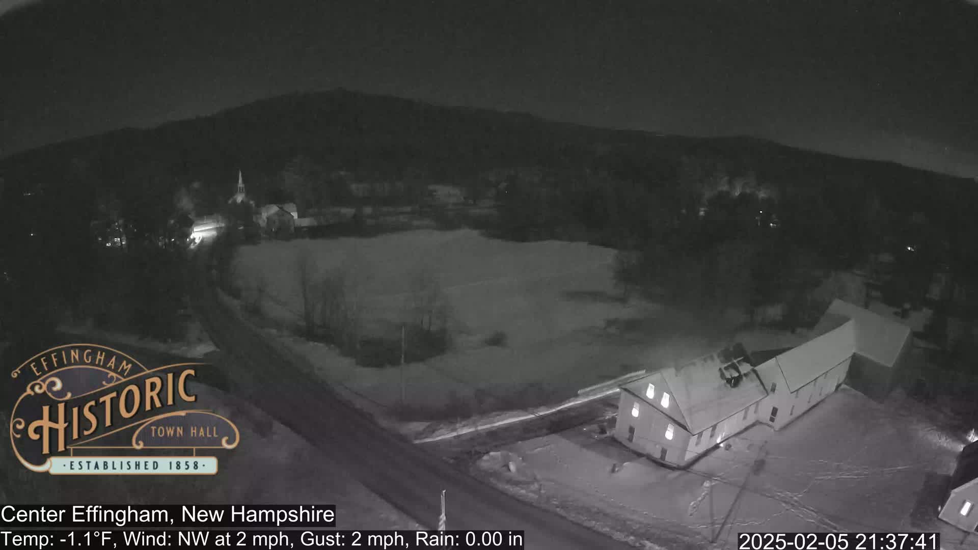 A nighttime, snow-covered landscape shows a small town with a building in the foreground under a clear sky.