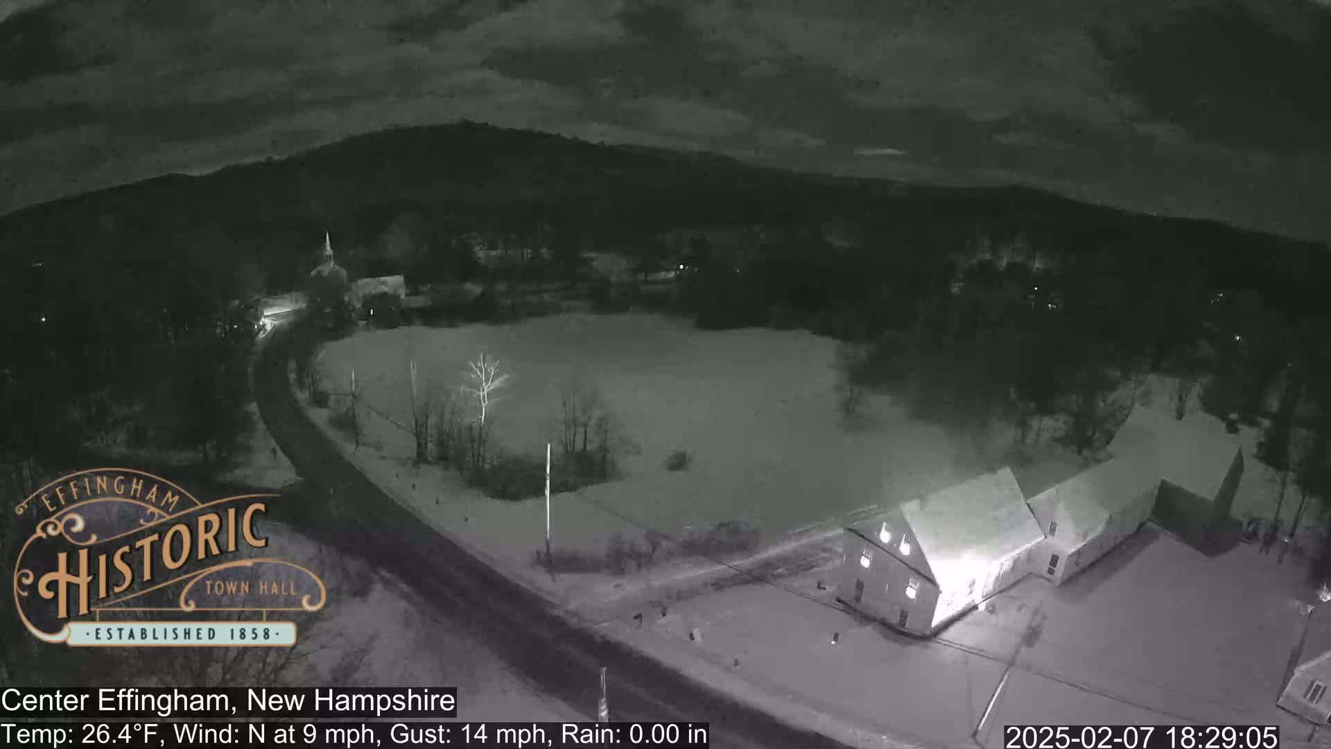 A nighttime, snow-covered scene shows a road curving past buildings and a field, under a dark sky.