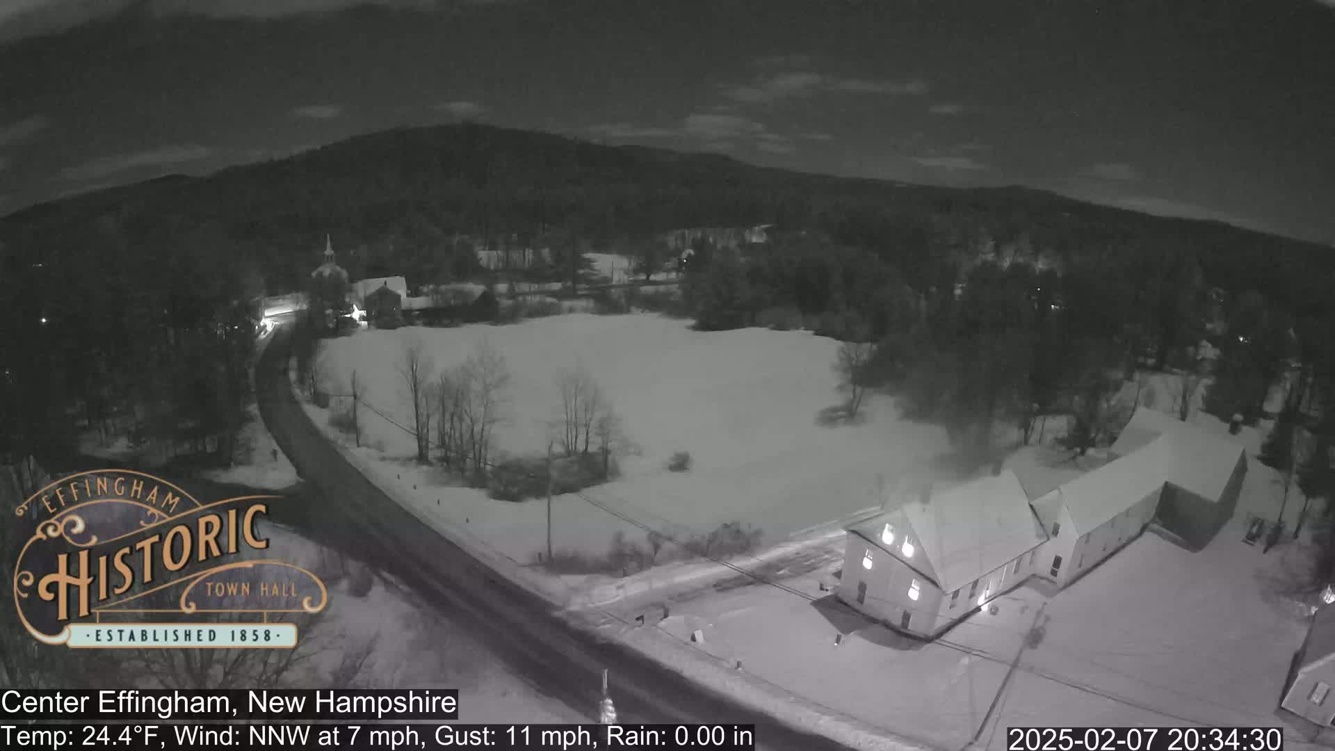 A snow-covered town at night, viewed from above, shows a road curving through a sparsely populated area with buildings and trees.