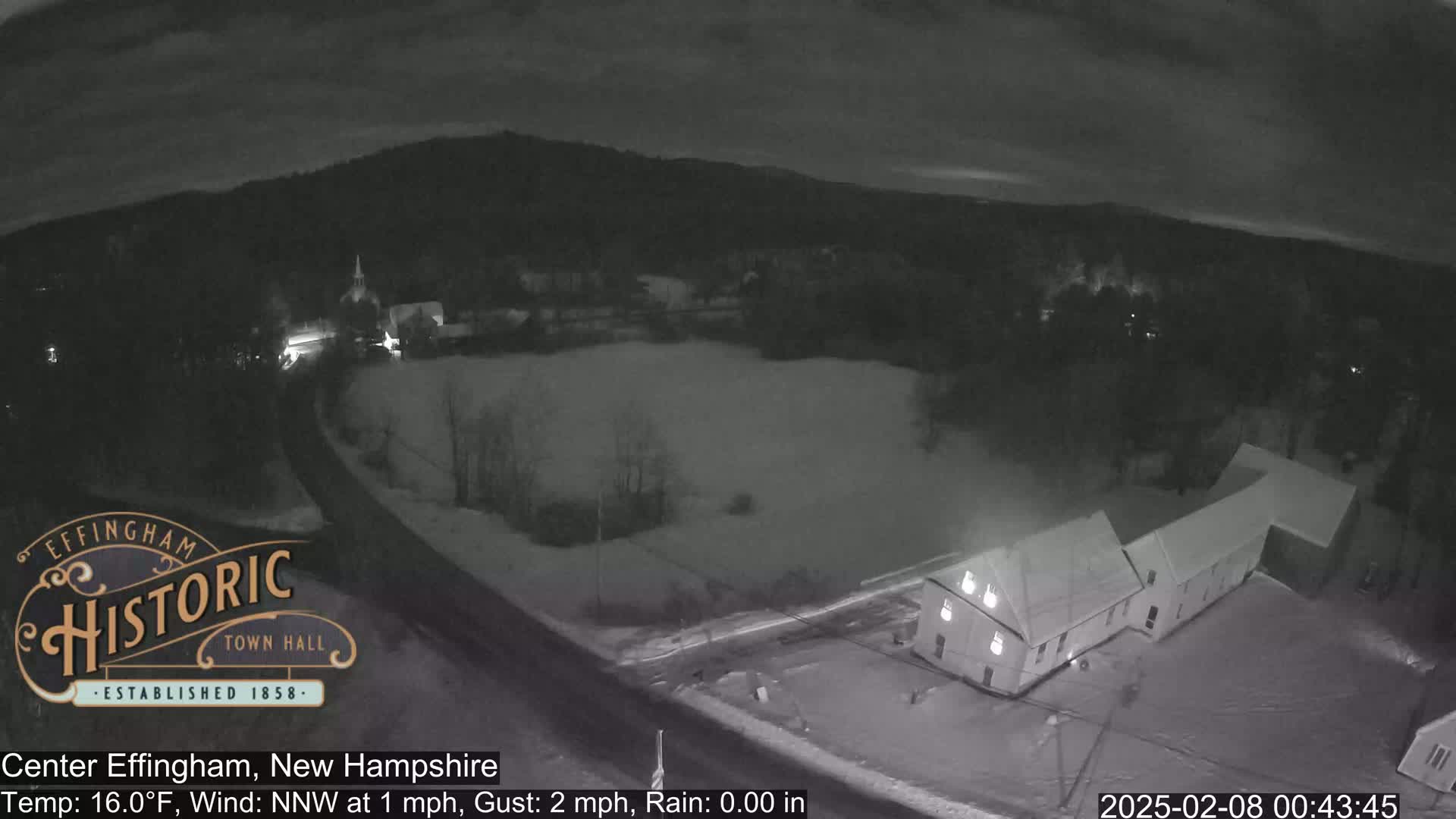 A nighttime, snowy scene shows a road, buildings, and a sparsely populated area under a cloudy sky.