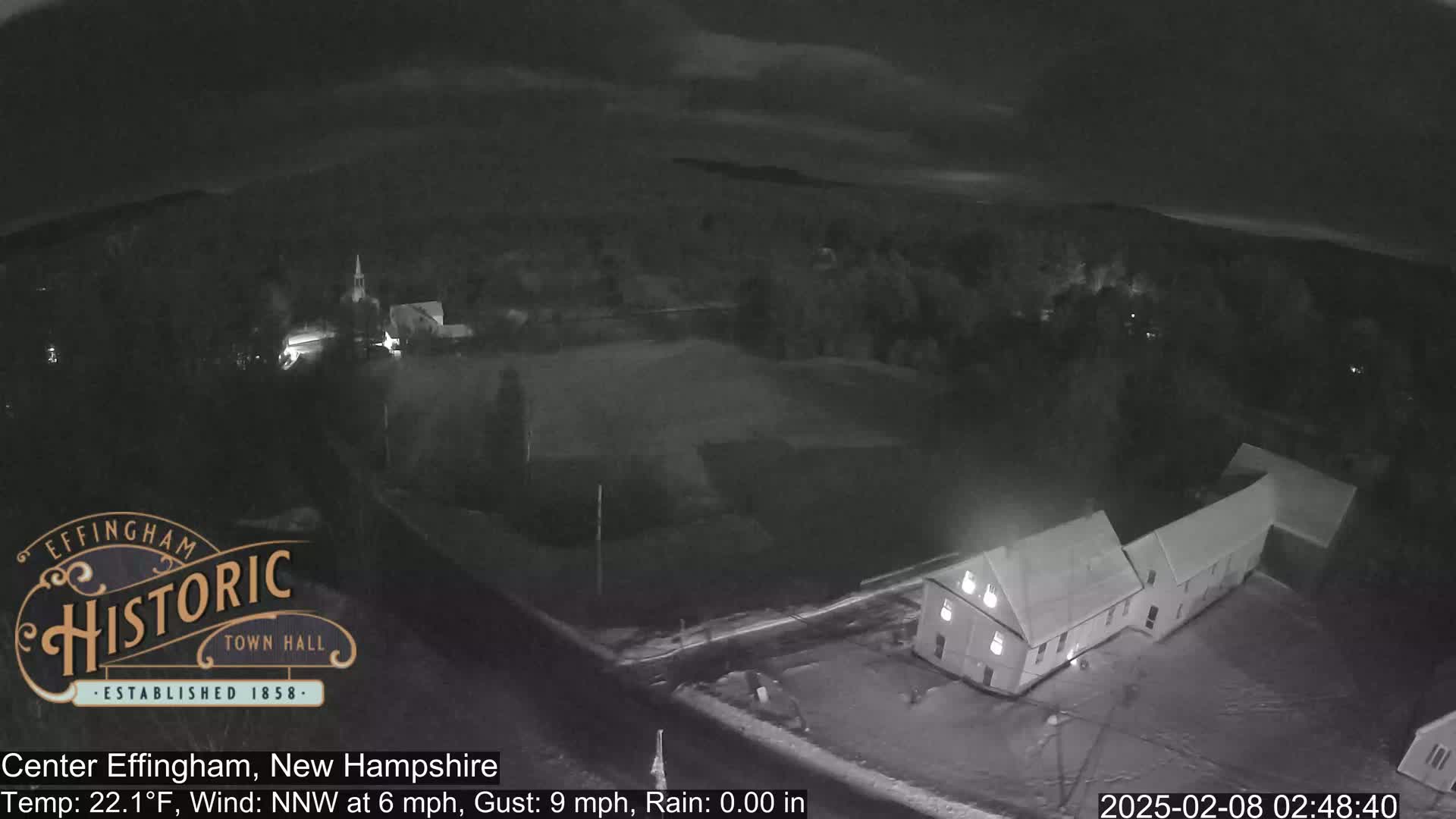 A nighttime, overhead view of a small town with a building illuminated under a partly cloudy sky, with a light wind and no rain.
