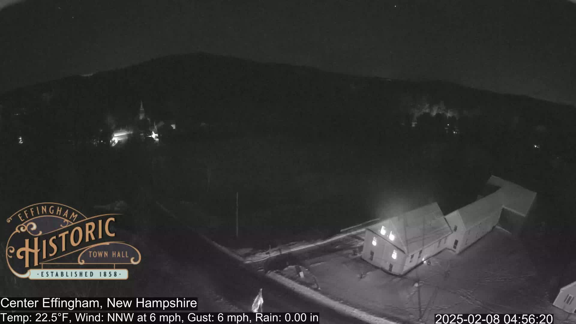A nighttime, snowy scene shows a building illuminated and a small town in the distance under a clear sky.