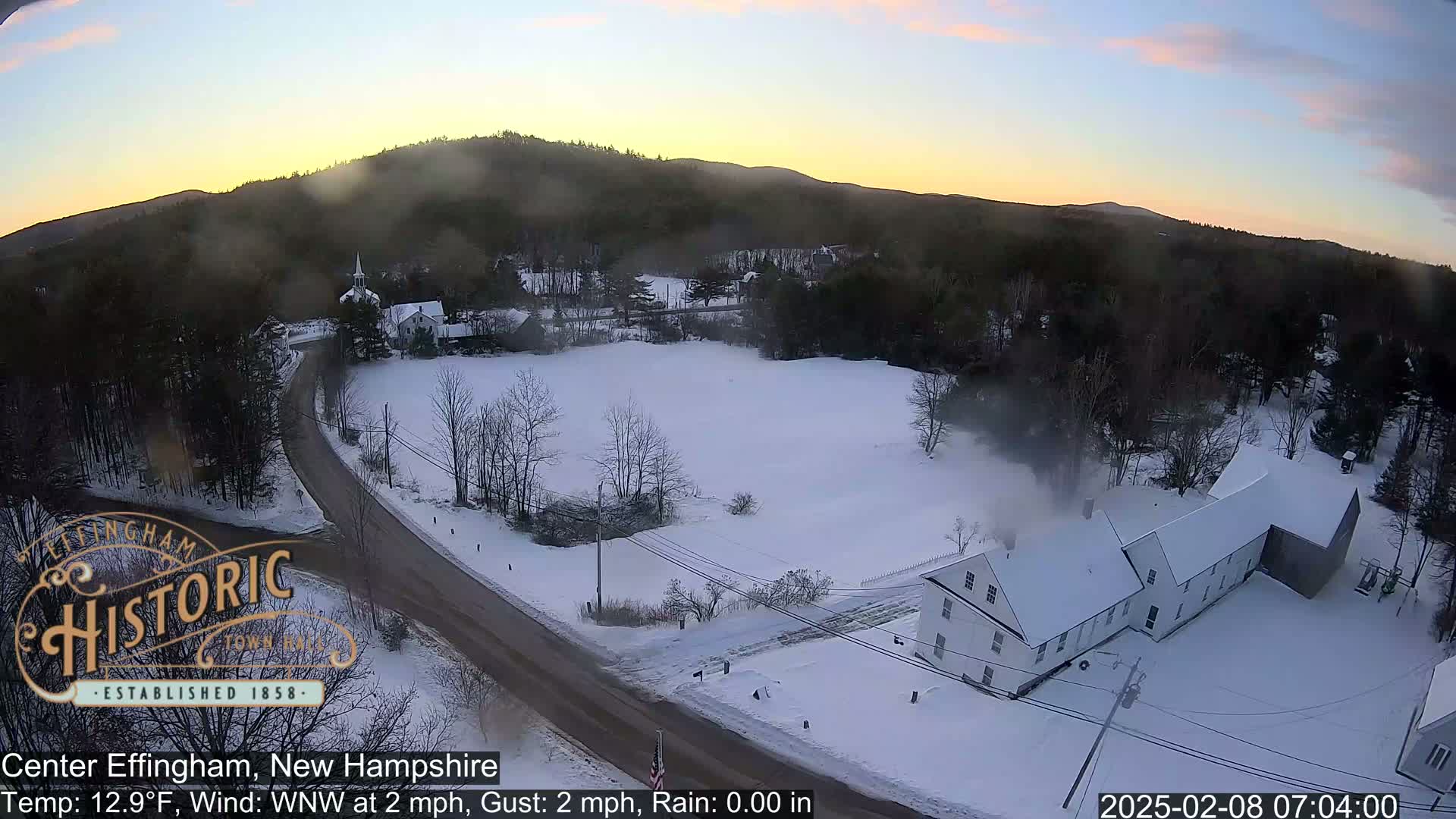 A snow-covered rural landscape at sunrise, showing a road, buildings, and trees, with a light wind.