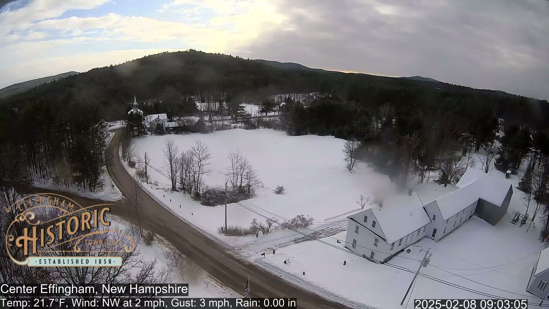 A snow-covered landscape shows a road, buildings, and a wooded area under an overcast sky.