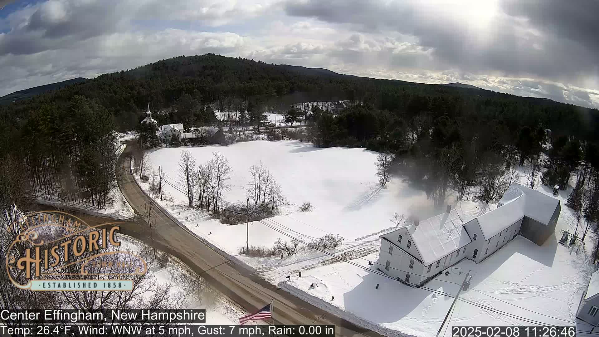 A snow-covered rural landscape with a road curving through it, featuring buildings and a forested hillside under a partly cloudy sky.