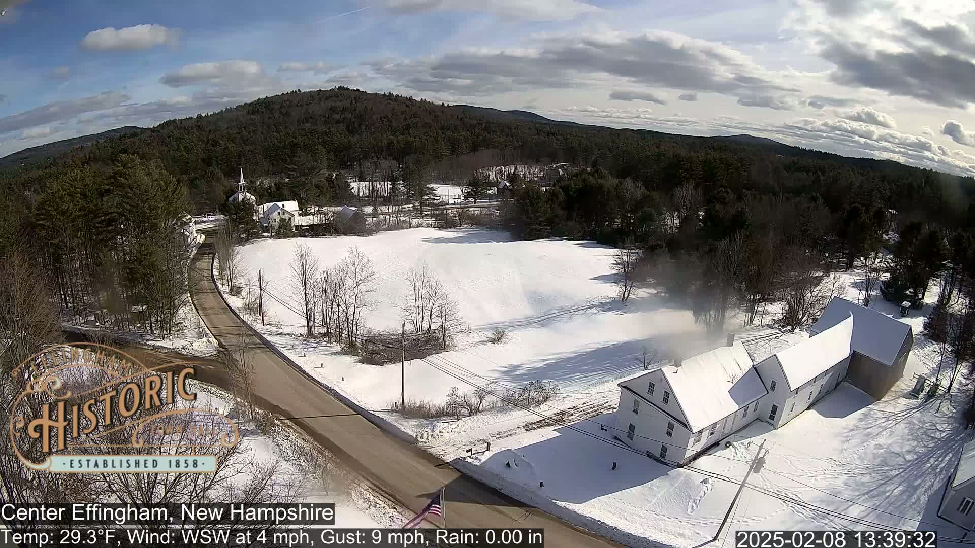 A snow-covered road winds through a village nestled among snow-covered trees and hills under a partly cloudy sky.