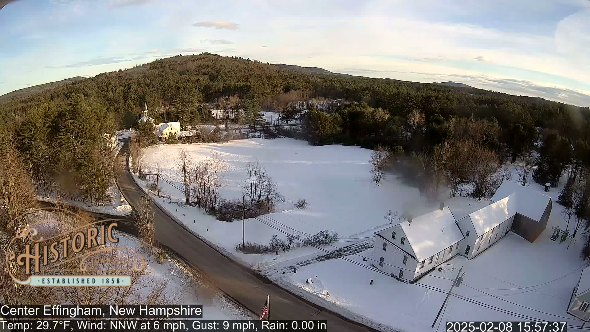 A snow-covered rural landscape with a road, houses, and a forested hillside under a mostly clear sky.
