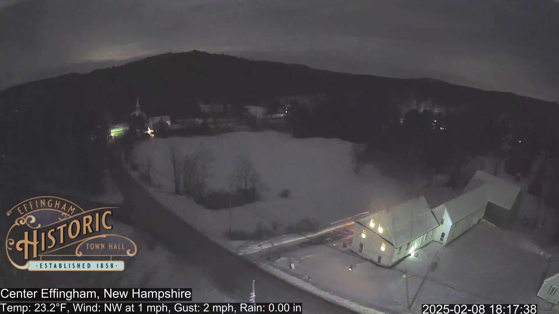 A nighttime, snow-covered landscape with a road, buildings, and trees under a dark sky.