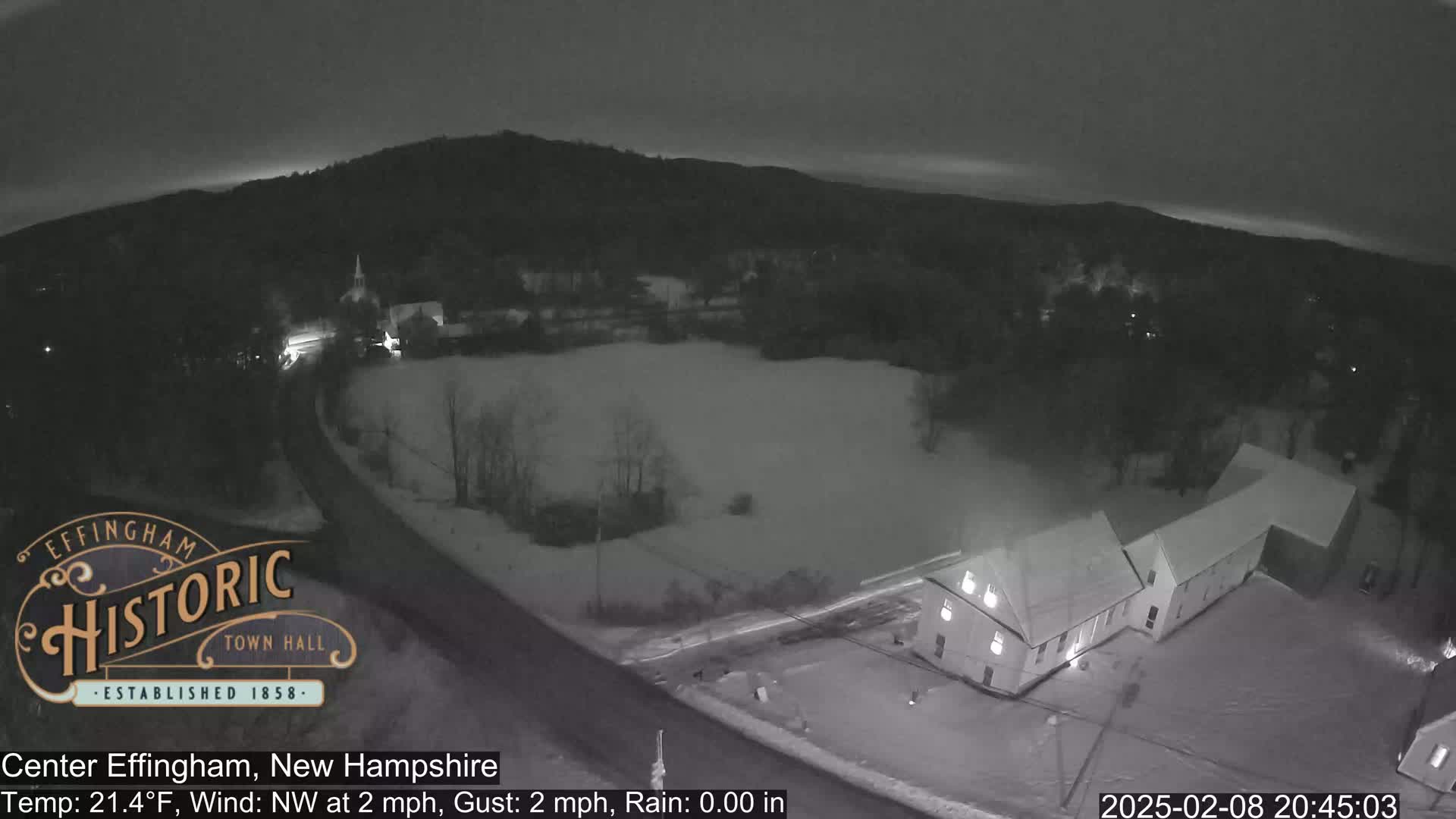 A nighttime, snow-covered scene shows a road curving past buildings and a field, under a dark sky with a northwest wind.