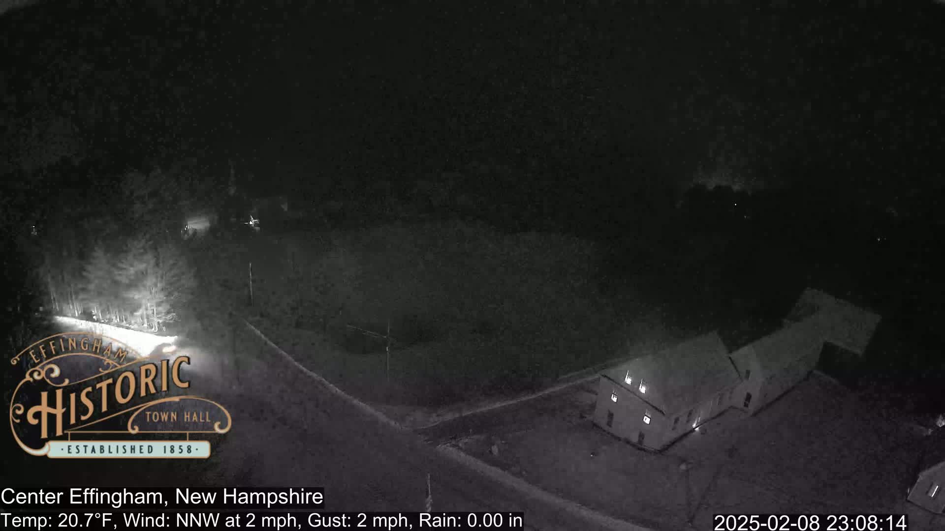 A nighttime, low-light view shows a road curving past a building and some trees, under a dark, clear sky with a 2 mph north-northwest wind and a temperature of 20.7°F.
