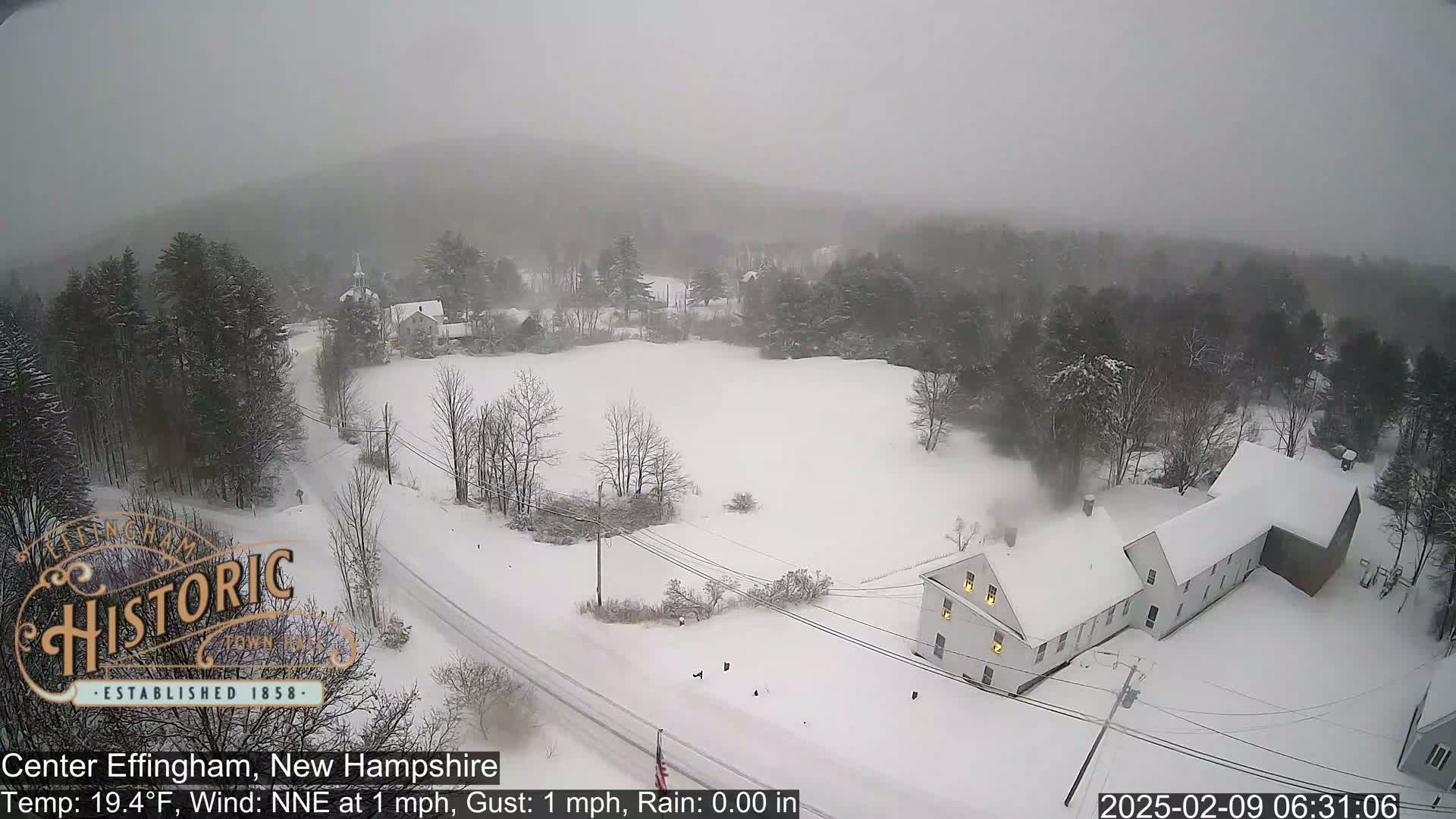 A snow-covered landscape shows a road and buildings under a gray, snowy sky.