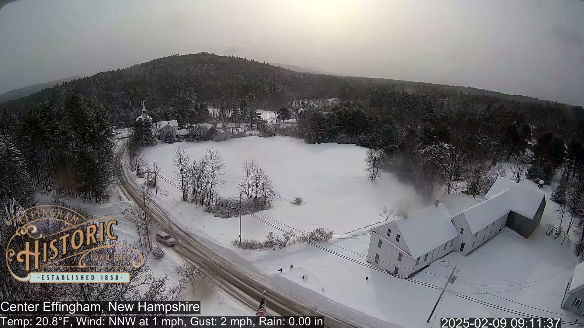 A snow-covered road winds through a snowy landscape, featuring a white building and a line of trees under a hazy sky.
