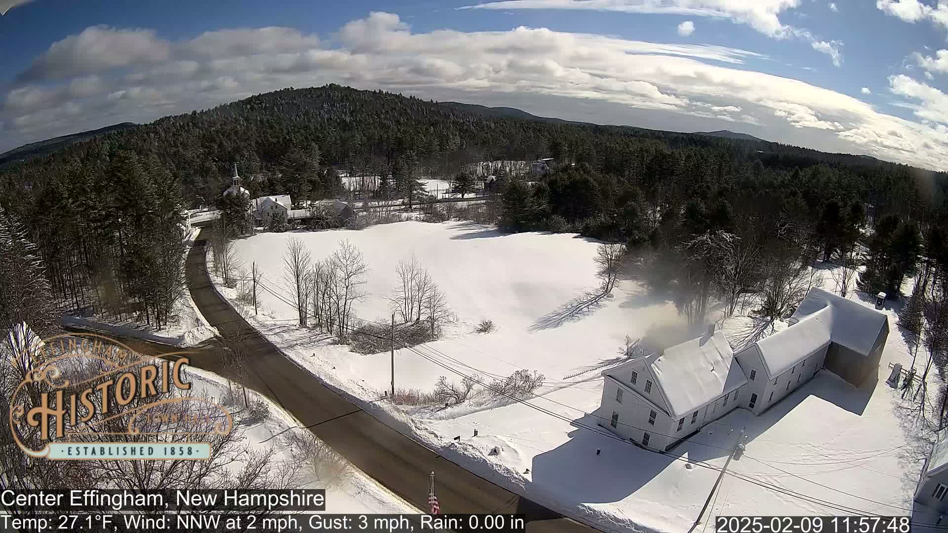 A snow-covered landscape with a road, buildings, and a forest under a partly cloudy sky.