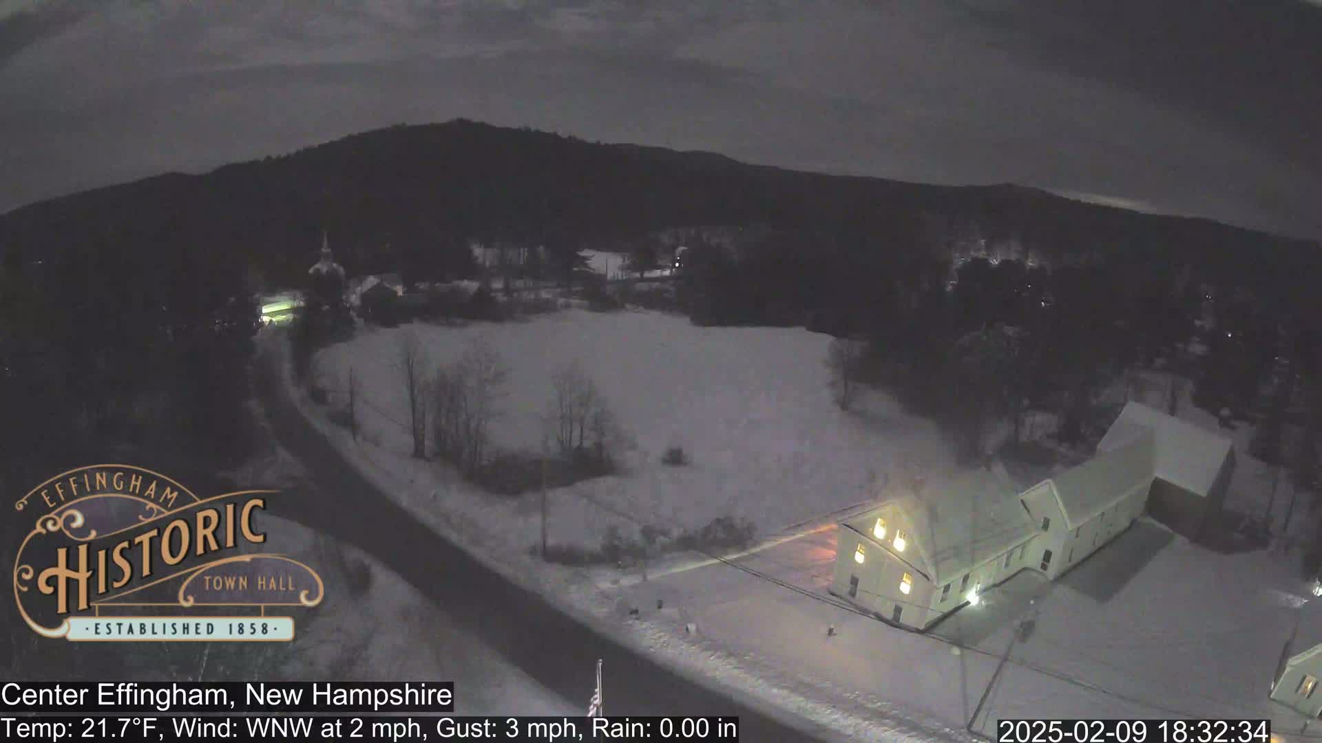 A snow-covered landscape at night, showing a road, buildings with illuminated windows, and a hill in the background under a dark sky.
