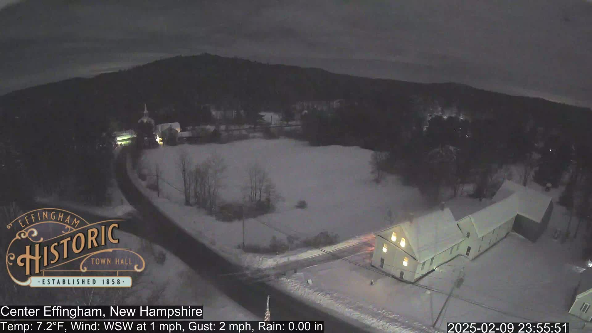 A snow-covered landscape at night is viewed from above, showing a road, buildings with lit windows, and a dark, snow-covered field.