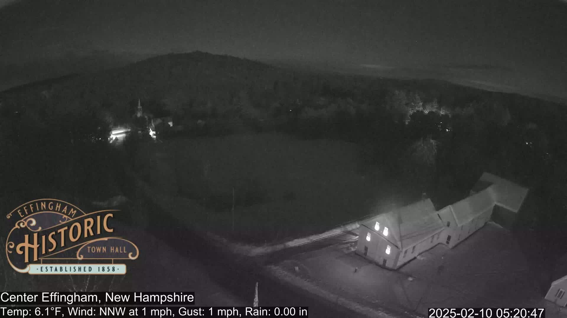 A nighttime, rainy view of a town with a long building, a field, and hills in the distance.
