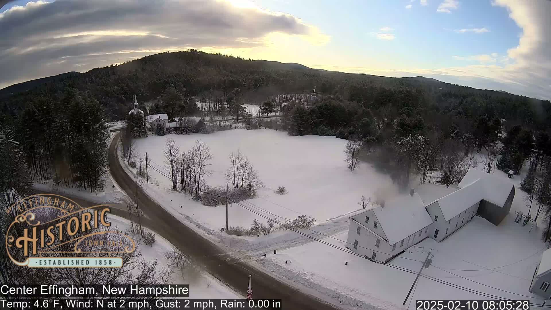 A snow-covered rural landscape with a road, buildings, and trees under a partly cloudy sky.