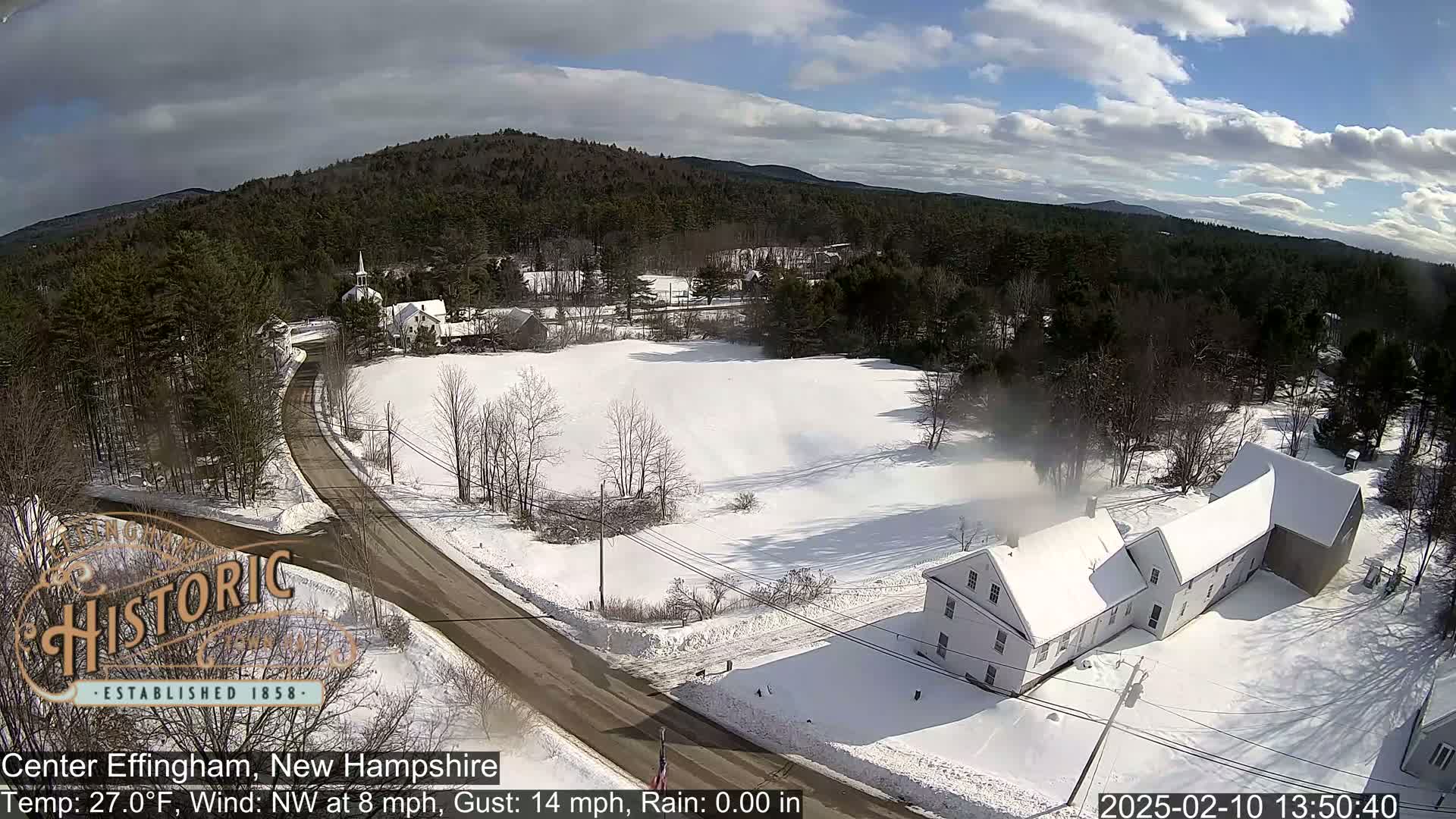 A snow-covered rural landscape with a road, buildings, and trees under partly sunny skies.