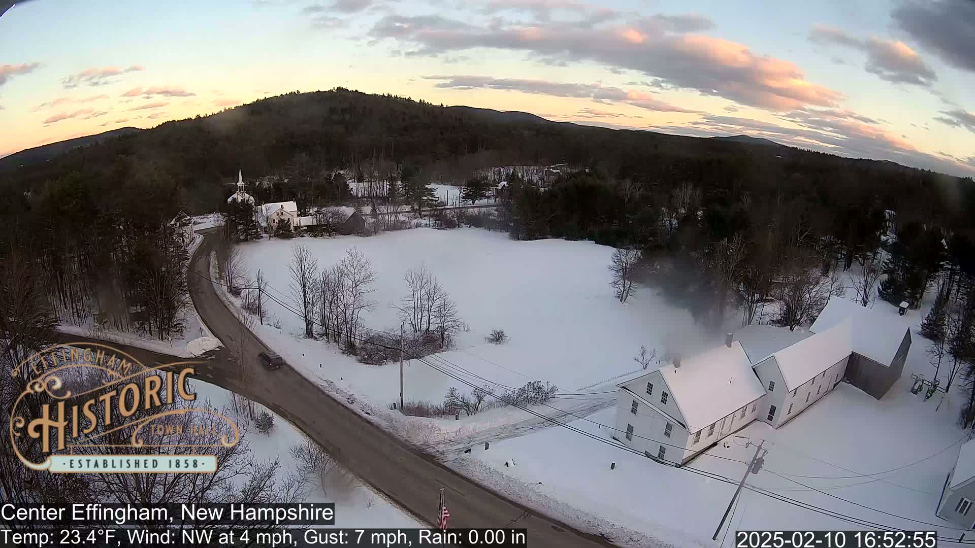 A snow-covered rural landscape at sunset, featuring a road, a church, several buildings, and a car, under partly cloudy skies.