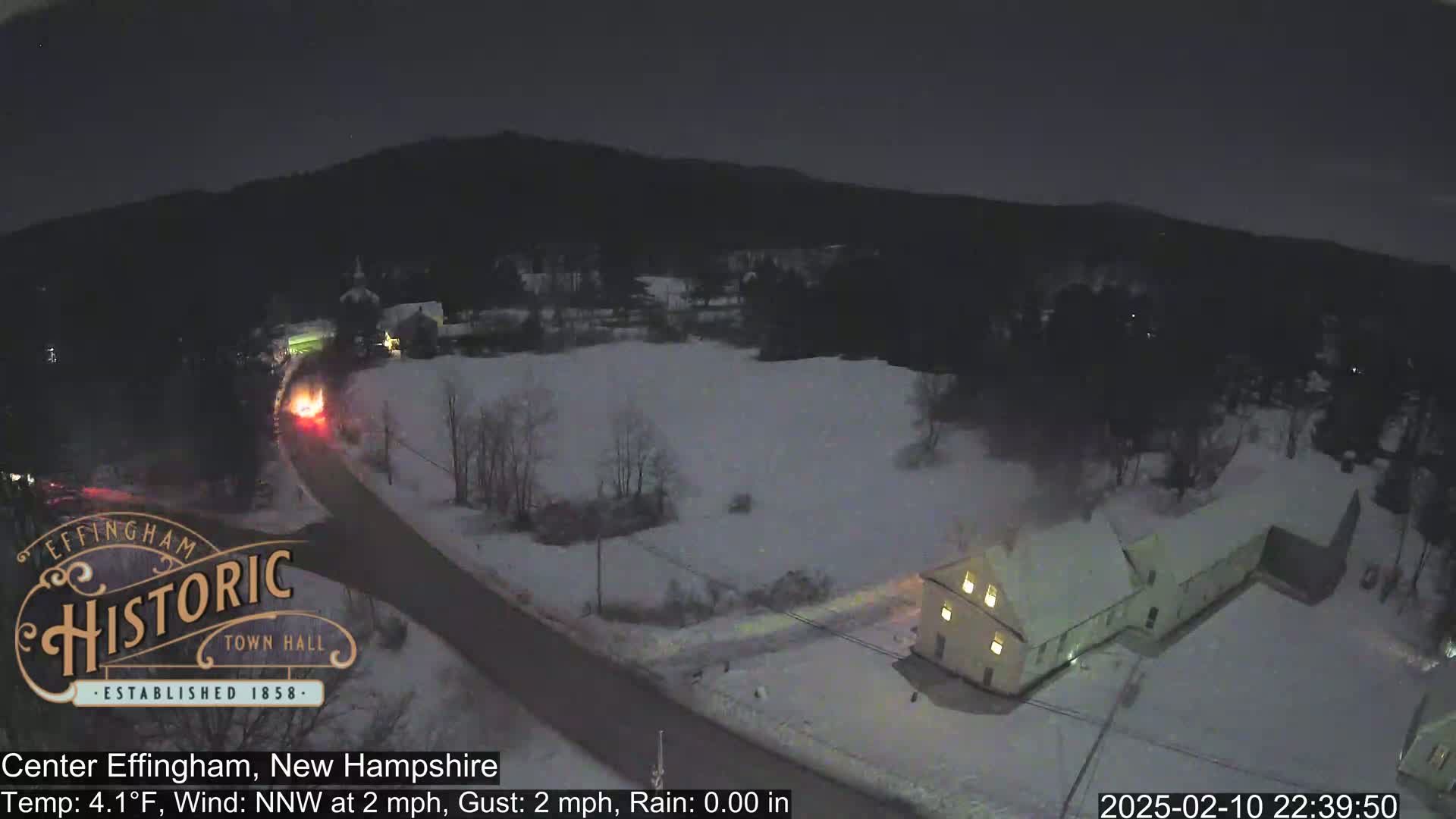 A nighttime aerial view of a snow-covered road and buildings in a small town, with a light rain.