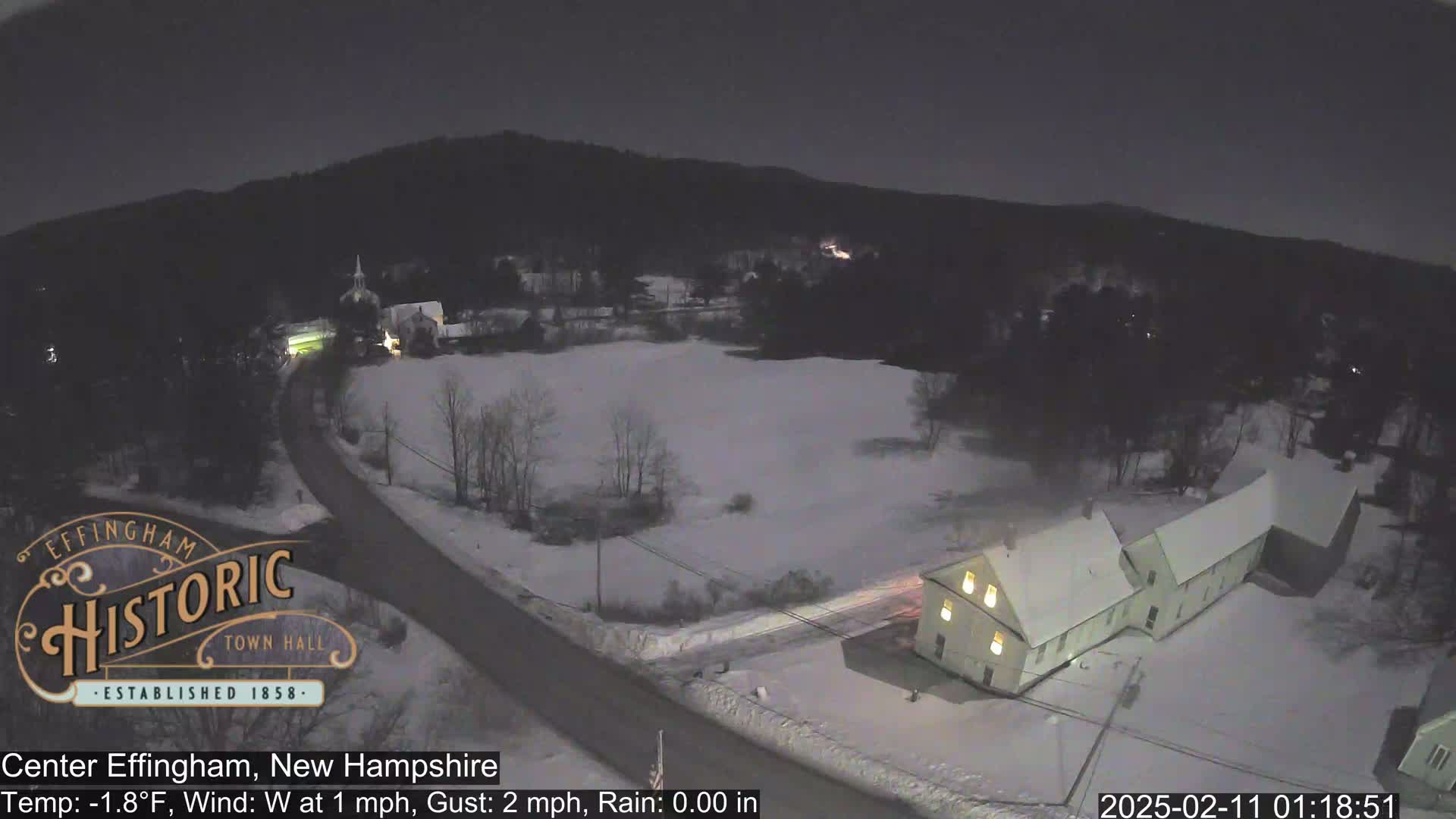 A snow-covered rural area at night, with a road curving through a field towards a building, under a dark sky.