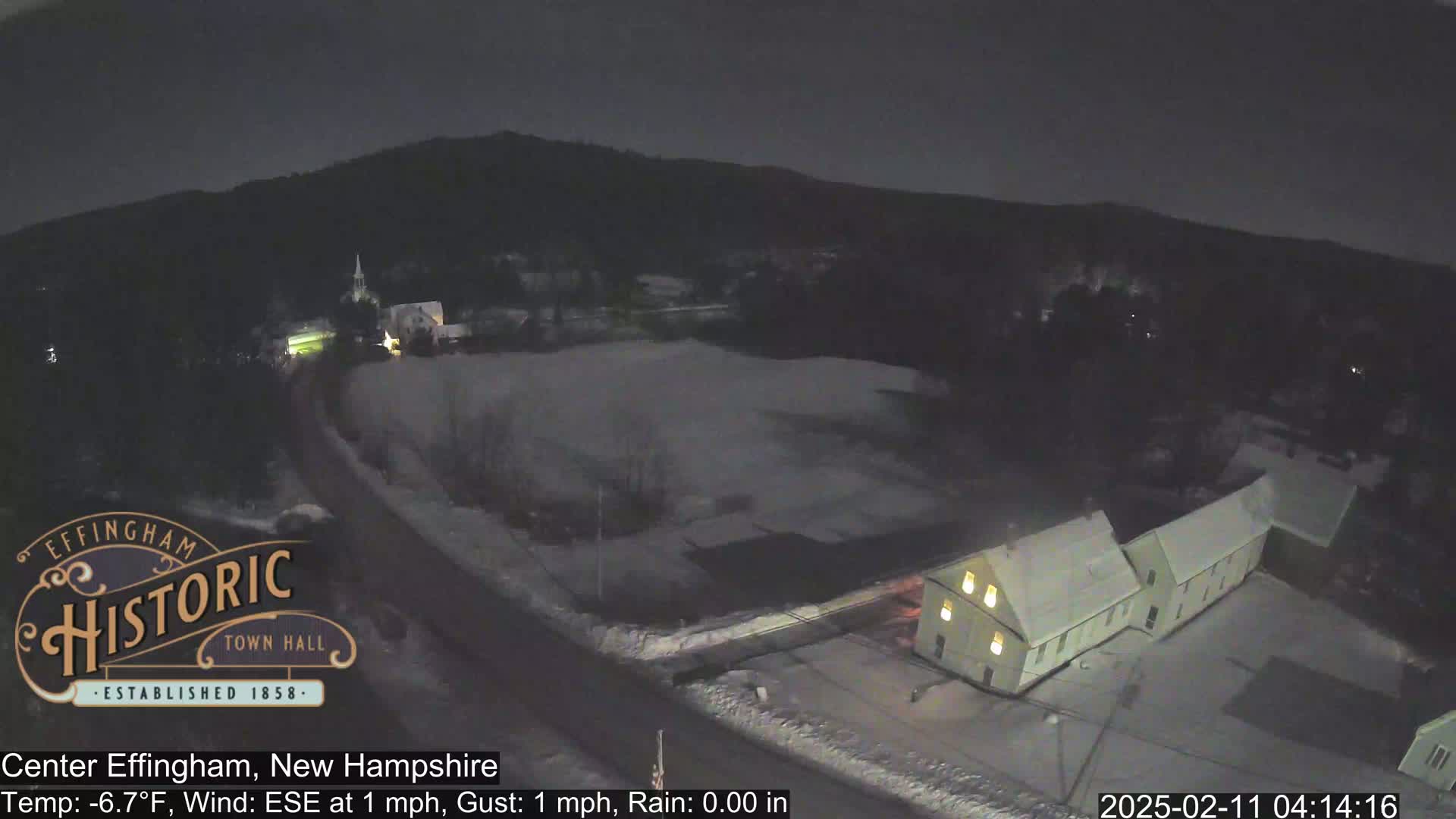 A nighttime, snowy scene shows a road winding past buildings under a dark sky.