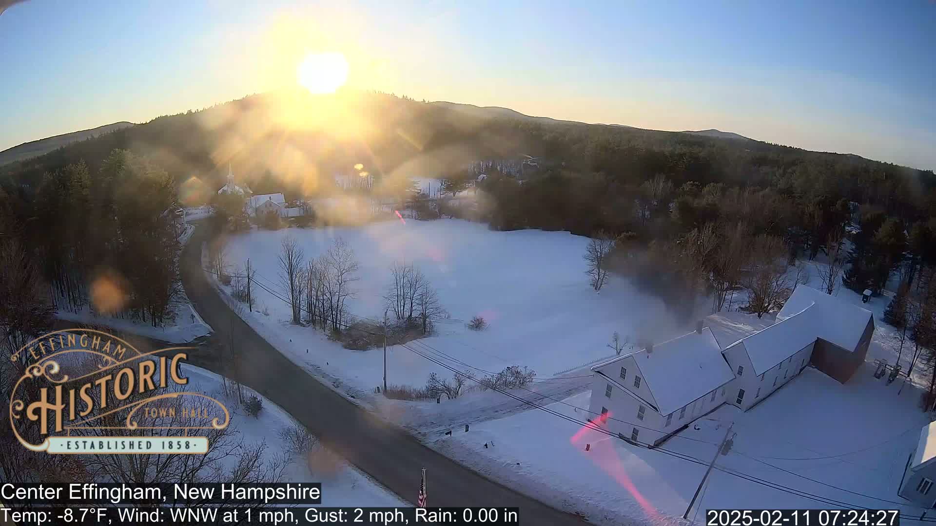 A snowy, sunlit scene shows a road curving through a snow-covered field toward a white building, with forested hills in the background.