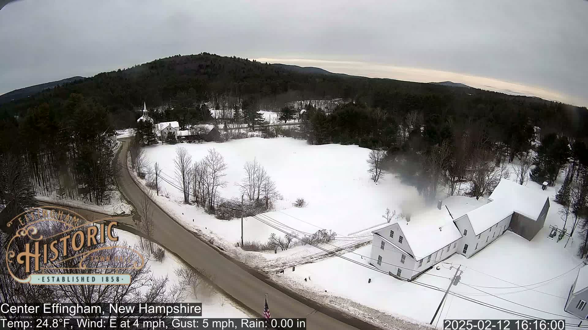 An aerial view of a snow-covered town in New Hampshire on an overcast day shows a road, buildings, and trees.