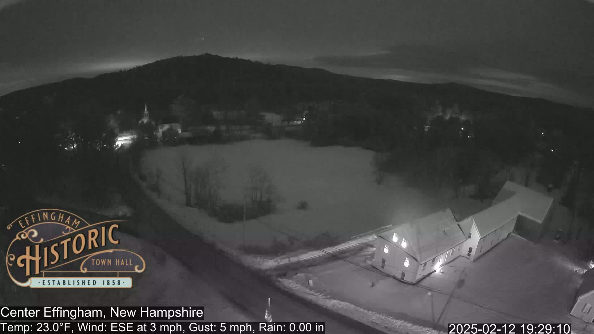 A nighttime, snow-covered scene shows a building, a road, and a field under a dark sky with a slight wind.