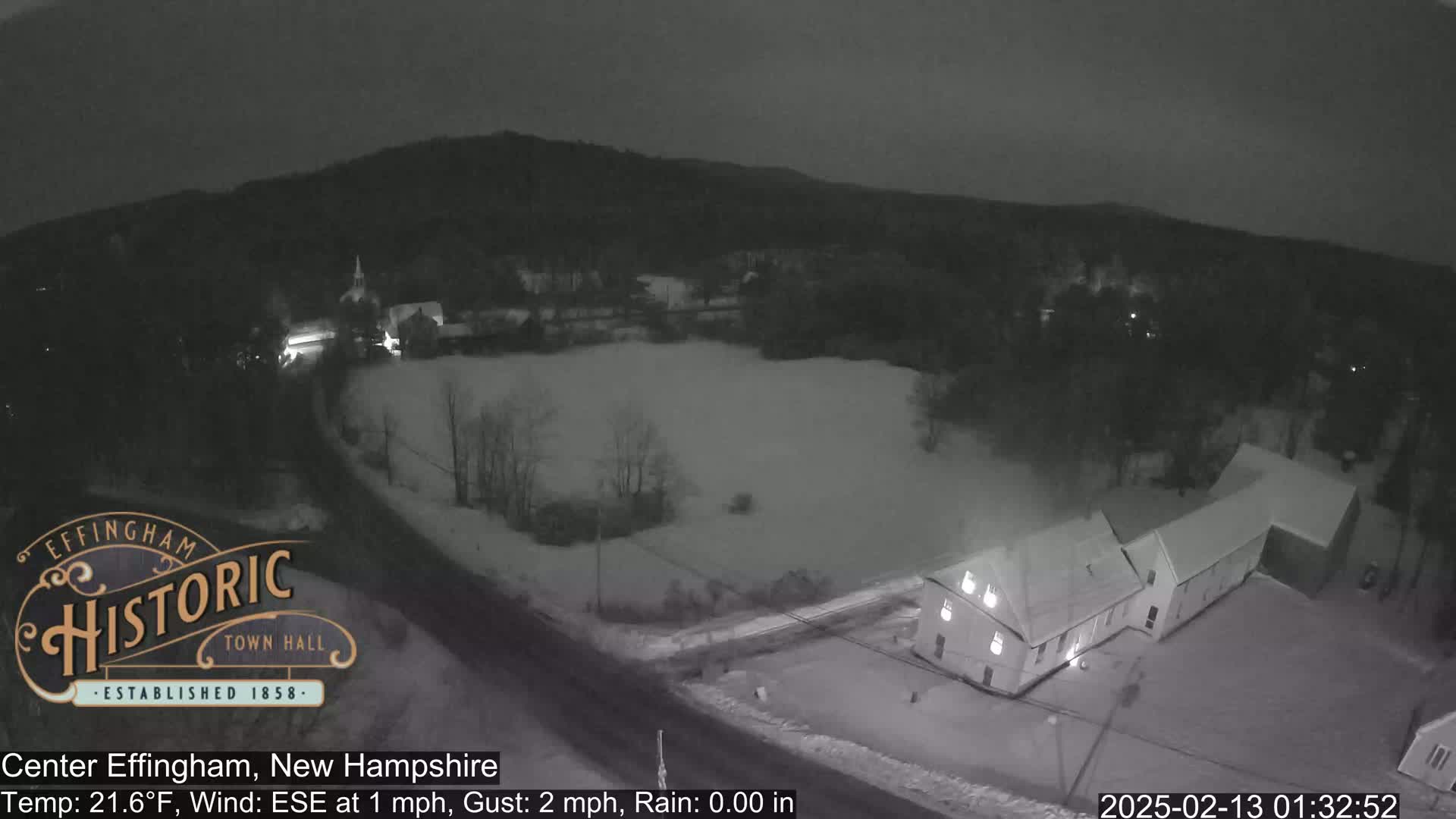A nighttime, snow-covered scene shows buildings and a road, with a slight wind from the east-southeast.