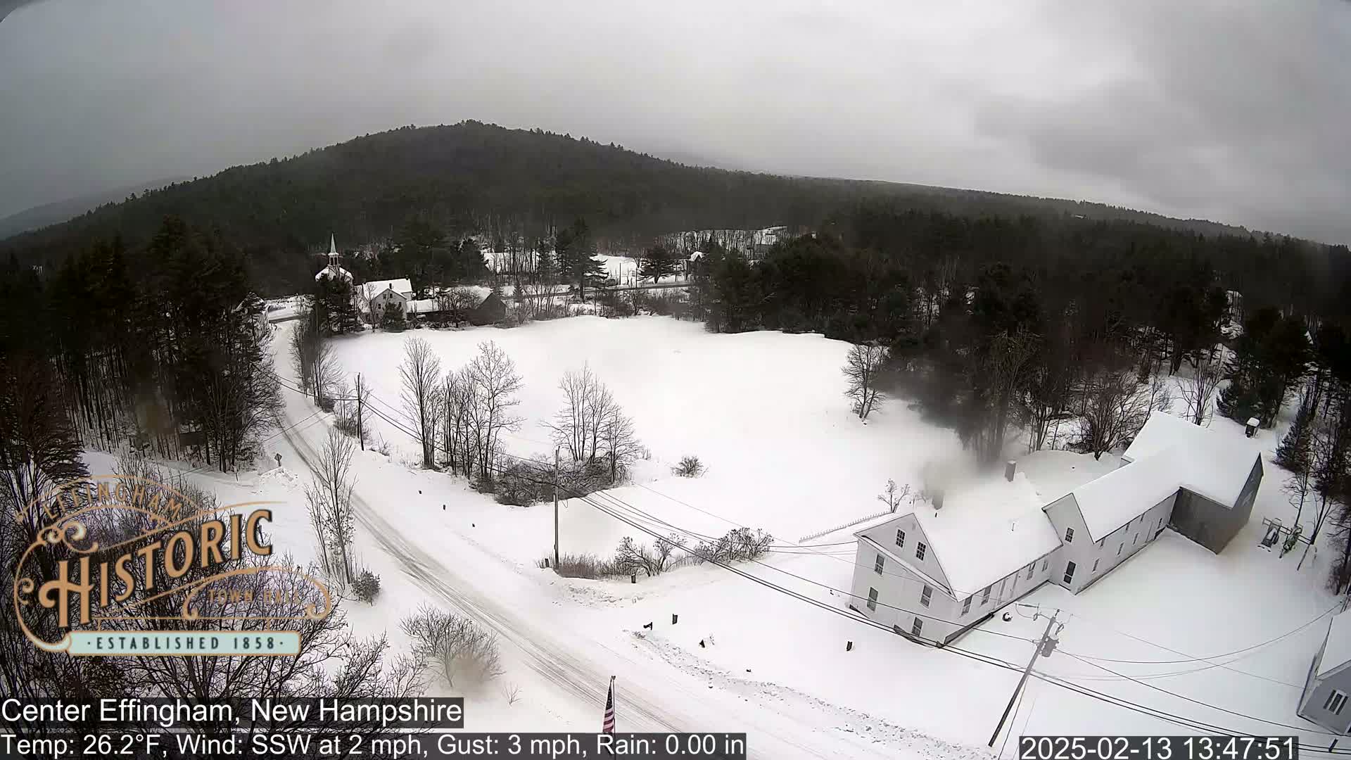 A snow-covered rural landscape with houses and a road, under a gray sky.