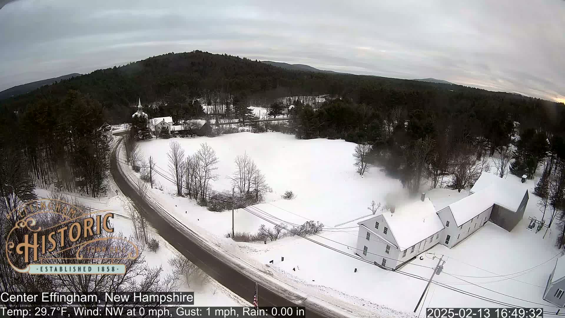 A snow-covered road curves through a rural area with several buildings and a snowy field, under an overcast sky.