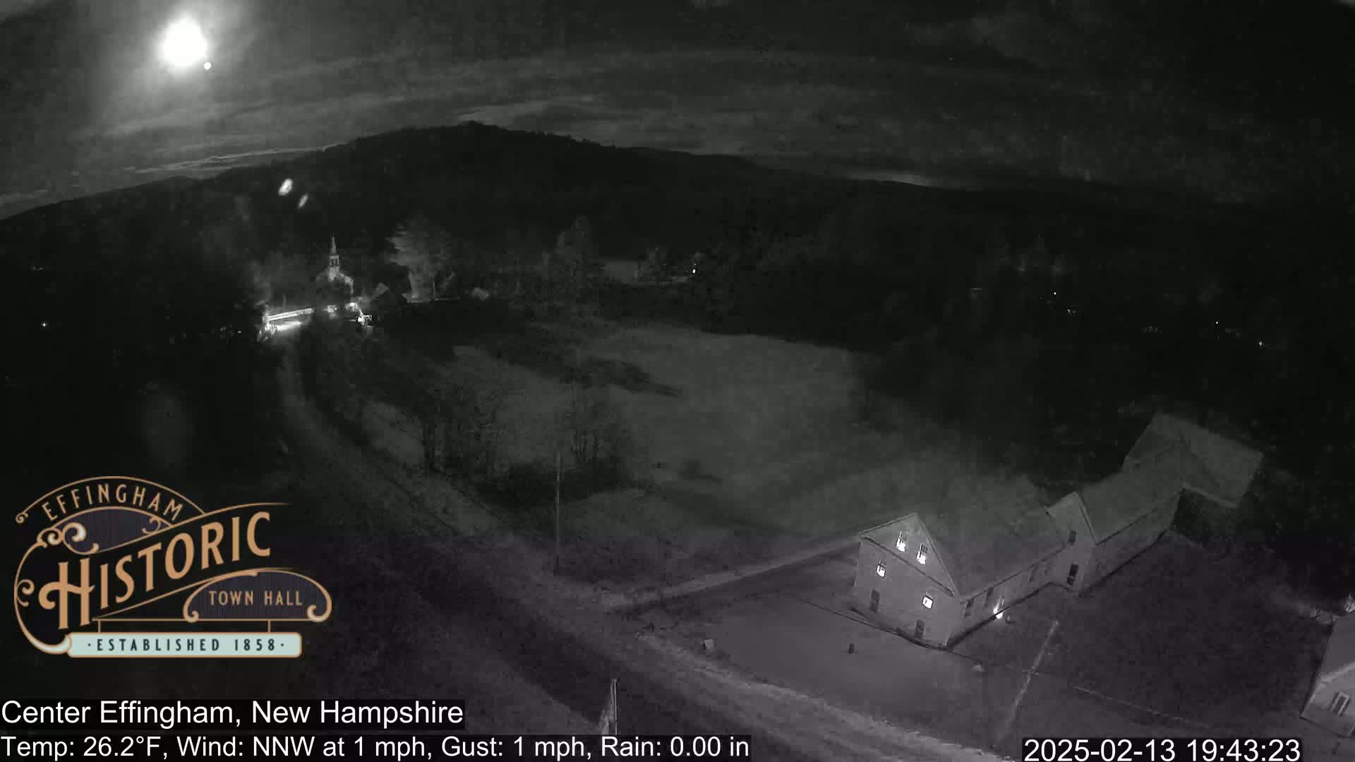 A nighttime, low-light view of a small town with a building and a road, under a partly cloudy sky with a bright moon.