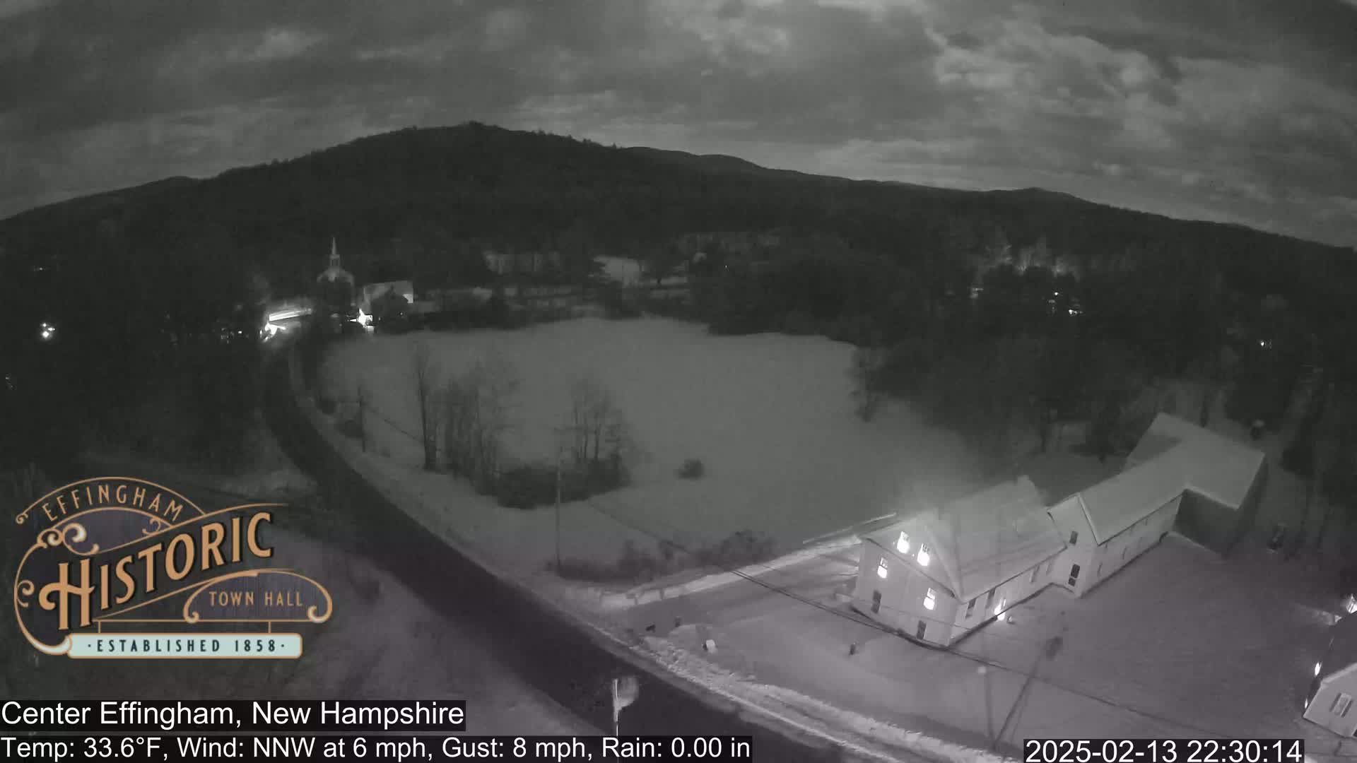 A nighttime, snowy scene shows a road, buildings, and a field, under a cloudy sky with a northwest wind.