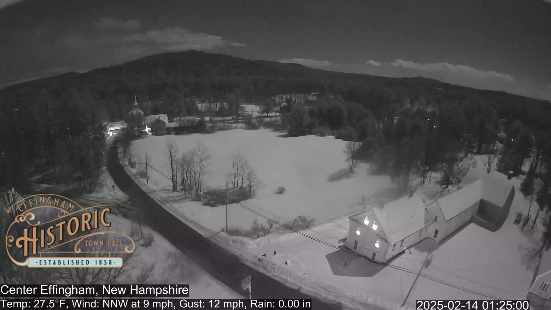 A nighttime aerial view of a snow-covered town with a road, buildings, and trees under a dark sky.