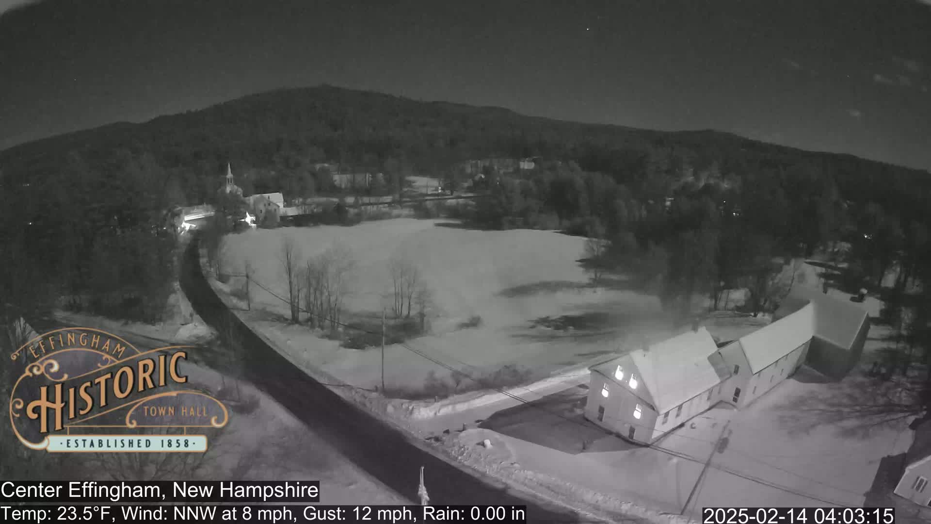 A nighttime, snow-covered landscape shows a road curving through a small town with buildings under a partly cloudy sky.