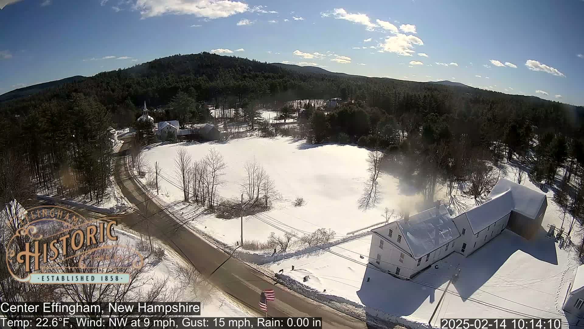 A snow-covered landscape shows a small town with buildings, roads, and trees under a mostly sunny sky.