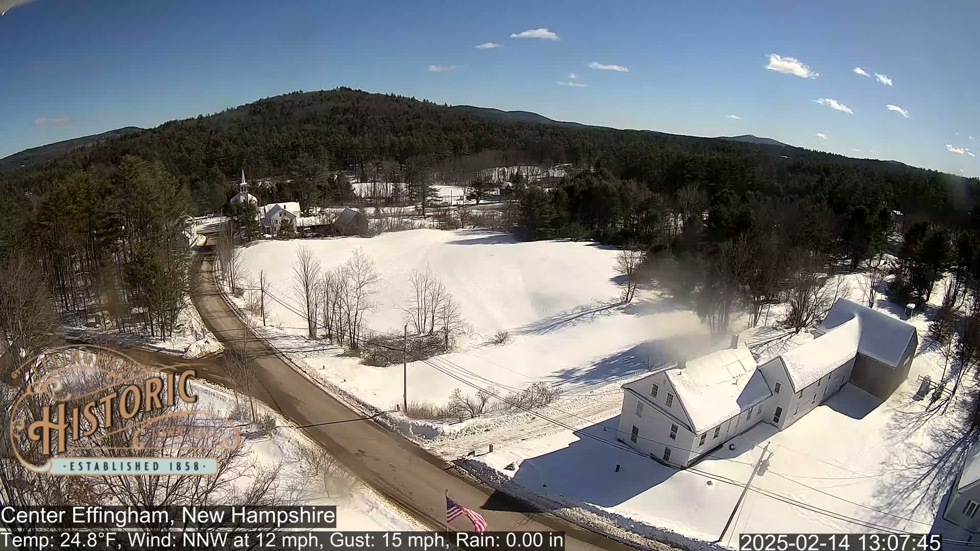 A snow-covered rural landscape with a road, houses, and trees under a clear, sunny sky.