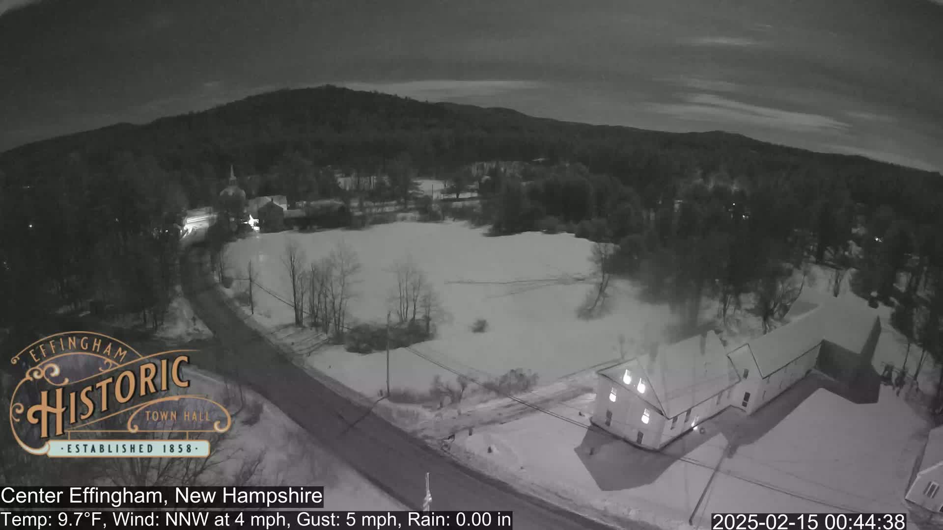 A nighttime aerial view shows a snow-covered town with a building near a road, under a dark, cloudy sky with light rain.