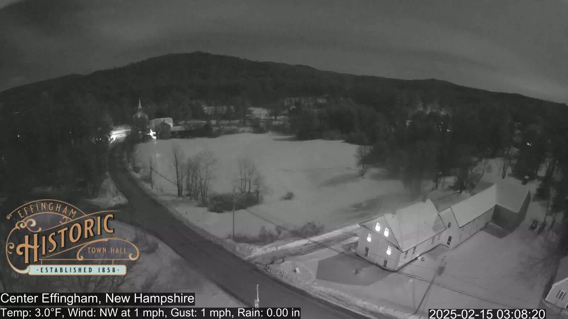 A nighttime aerial view of a snow-covered town with a road, buildings, and trees under a dark, cloudy sky.