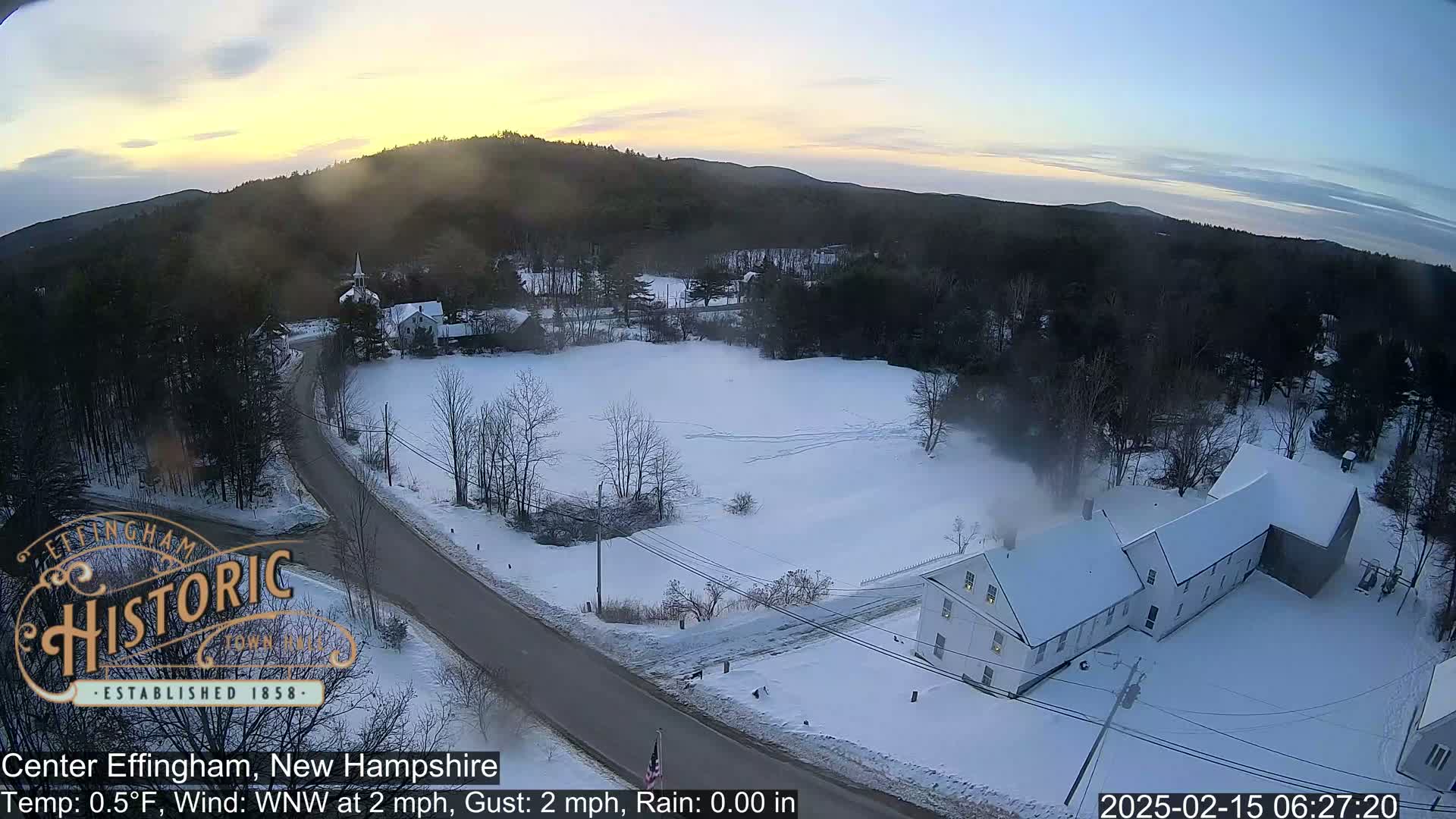 A snow-covered rural landscape at sunrise, featuring a road, buildings, and a field, with light winds.