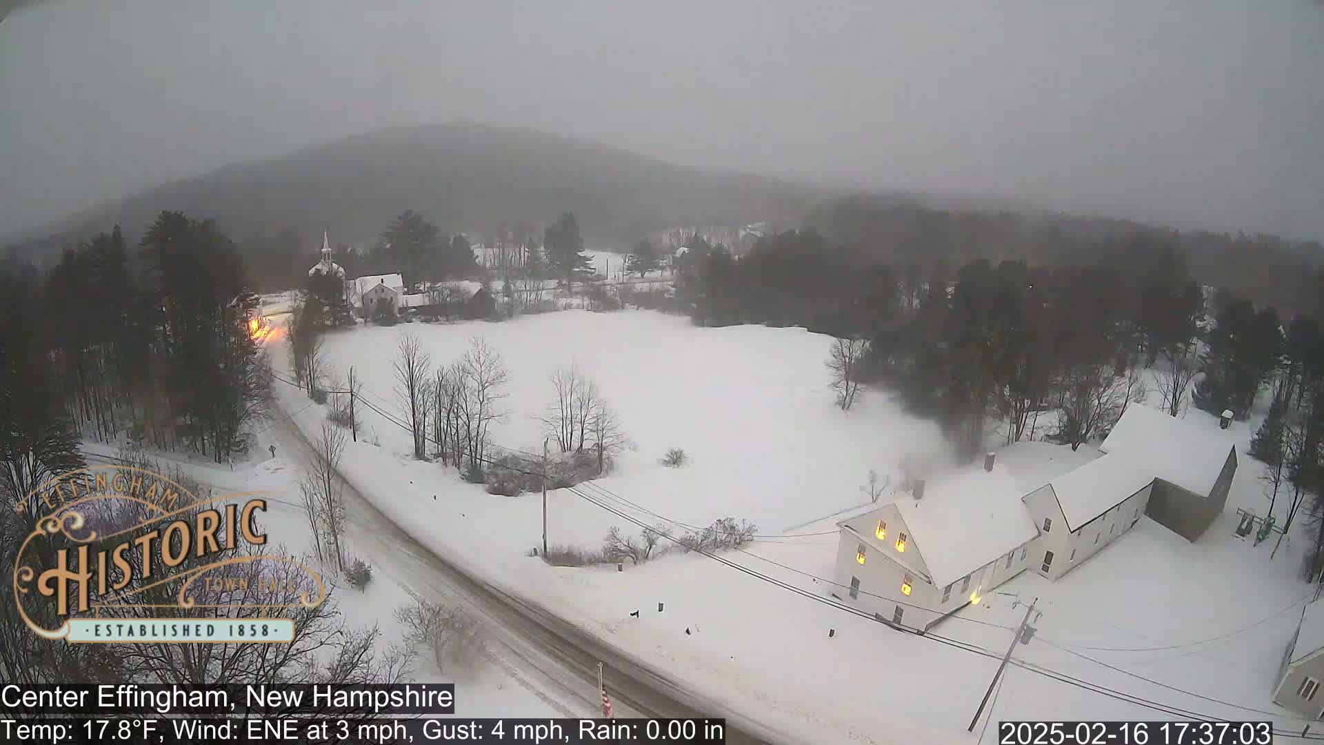 A snow-covered landscape with buildings and roads is seen under overcast skies during a light snowfall.