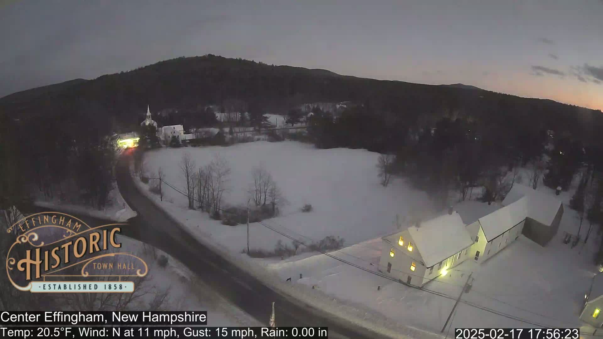 A snow-covered town at twilight, with a road and buildings visible under a dark, clear sky.