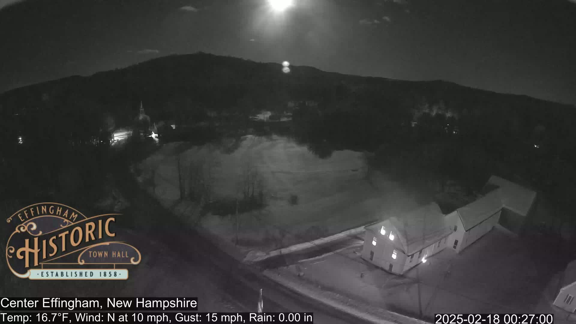 A nighttime, aerial view of a rural area shows a building illuminated against a dark landscape under a bright moon, with a slight north wind.