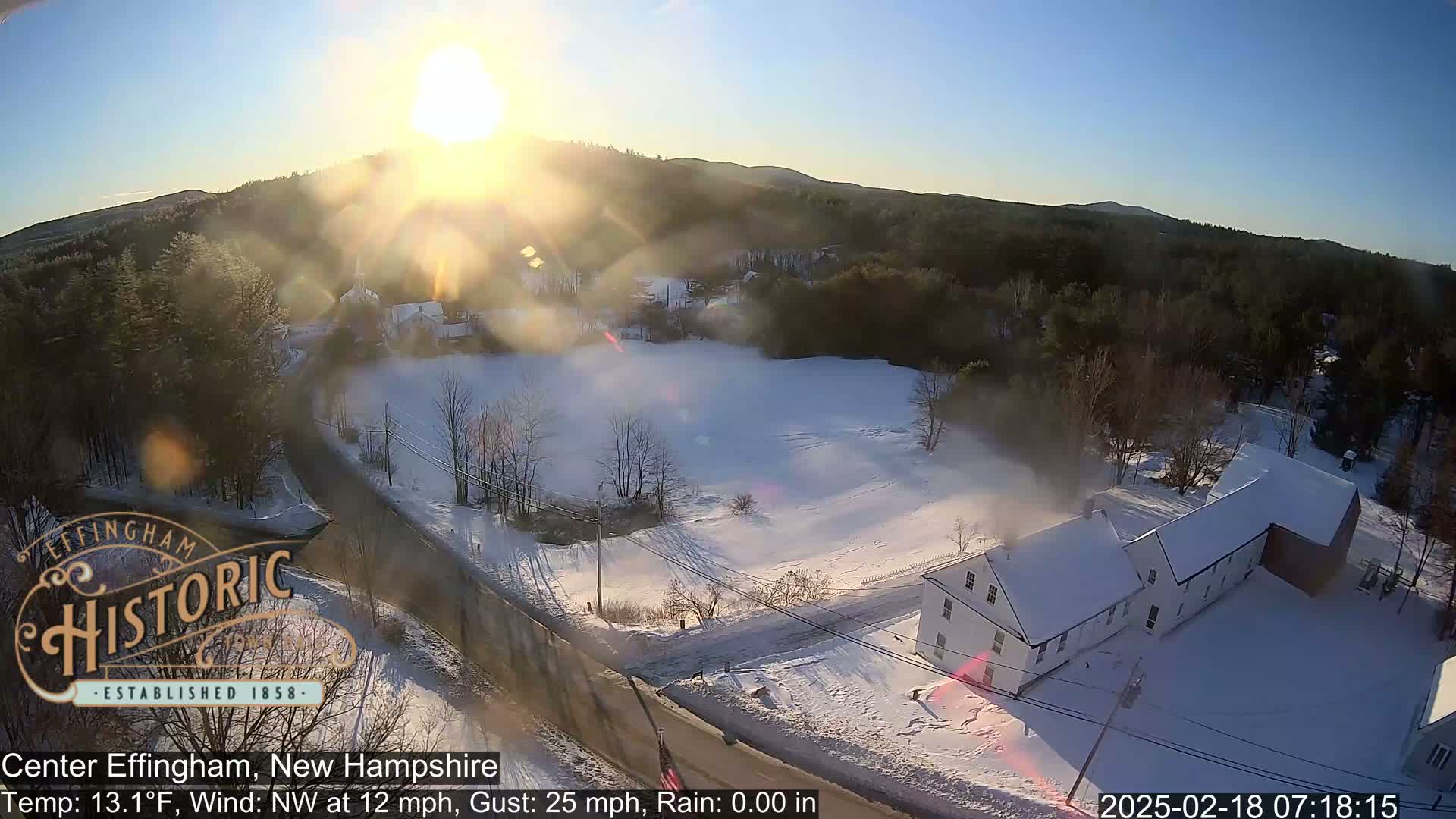 A snow-covered landscape with buildings and trees, viewed from above in bright, sunny conditions with a northwest wind.