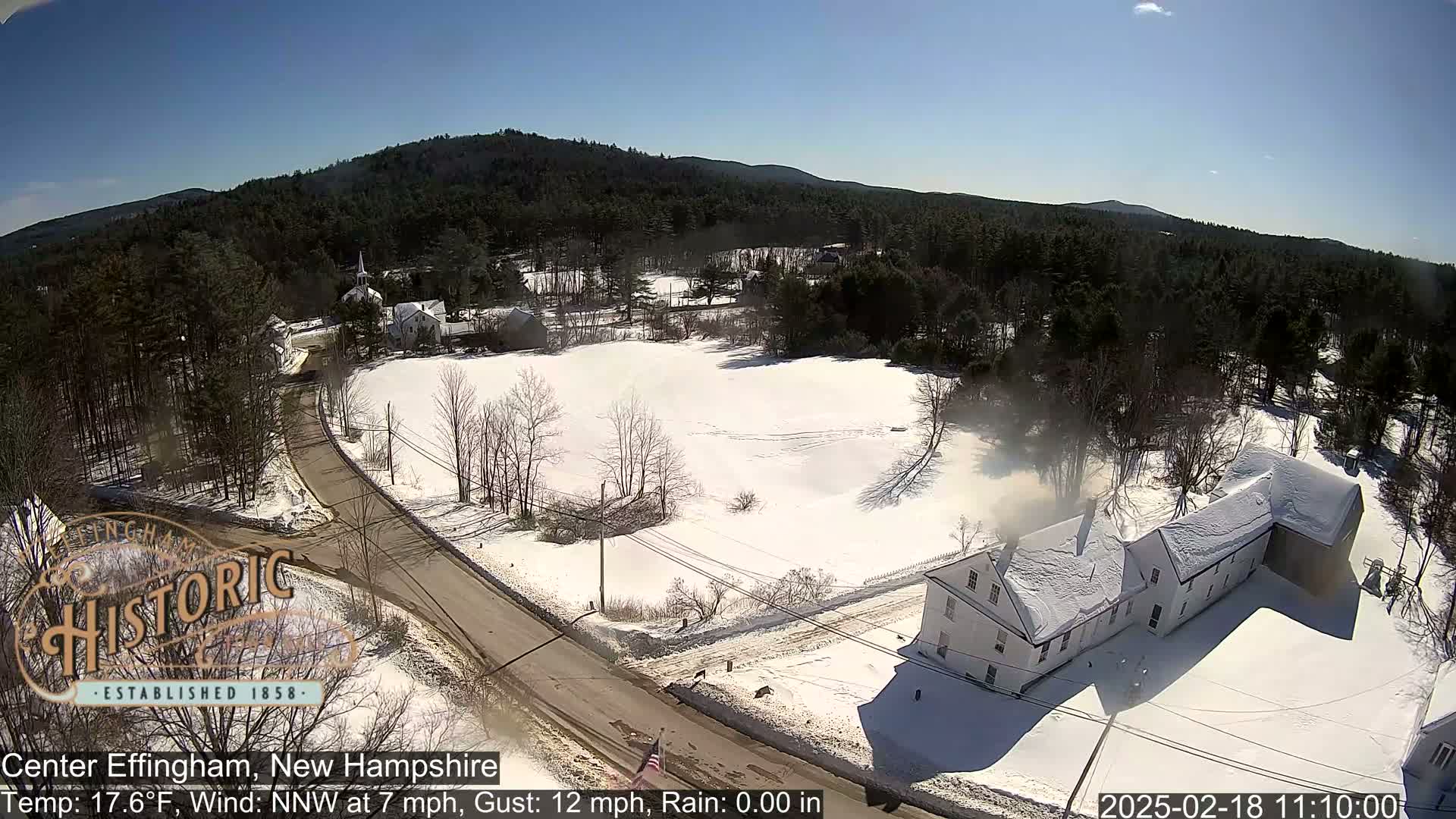 A snow-covered town, viewed from above, shows a road winding through a field and past buildings, under a clear blue sky.