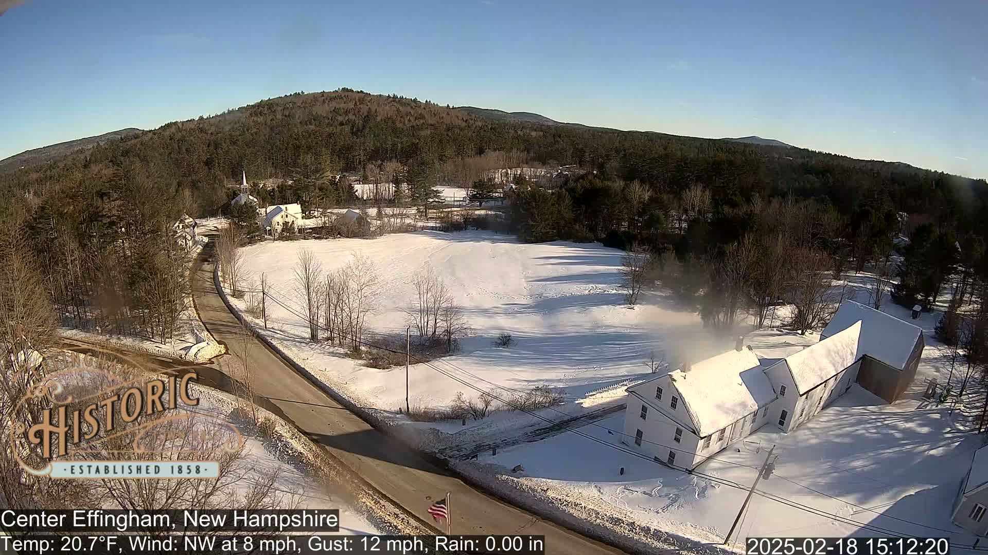 A snow-covered rural landscape with a road, several buildings, and a wooded hillside under a clear, sunny sky.