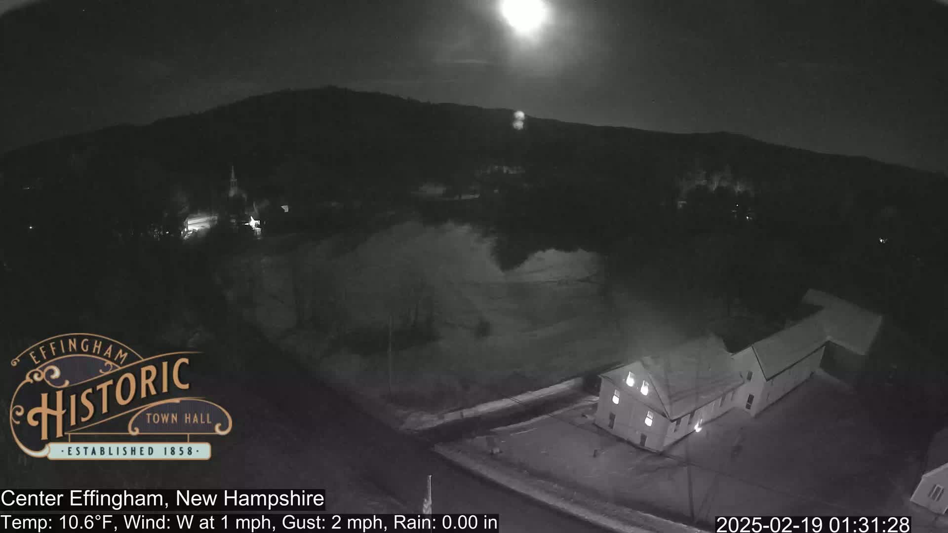 A nighttime, high-angle view of a small town under a bright moon, with a building illuminated in the foreground and light rain.