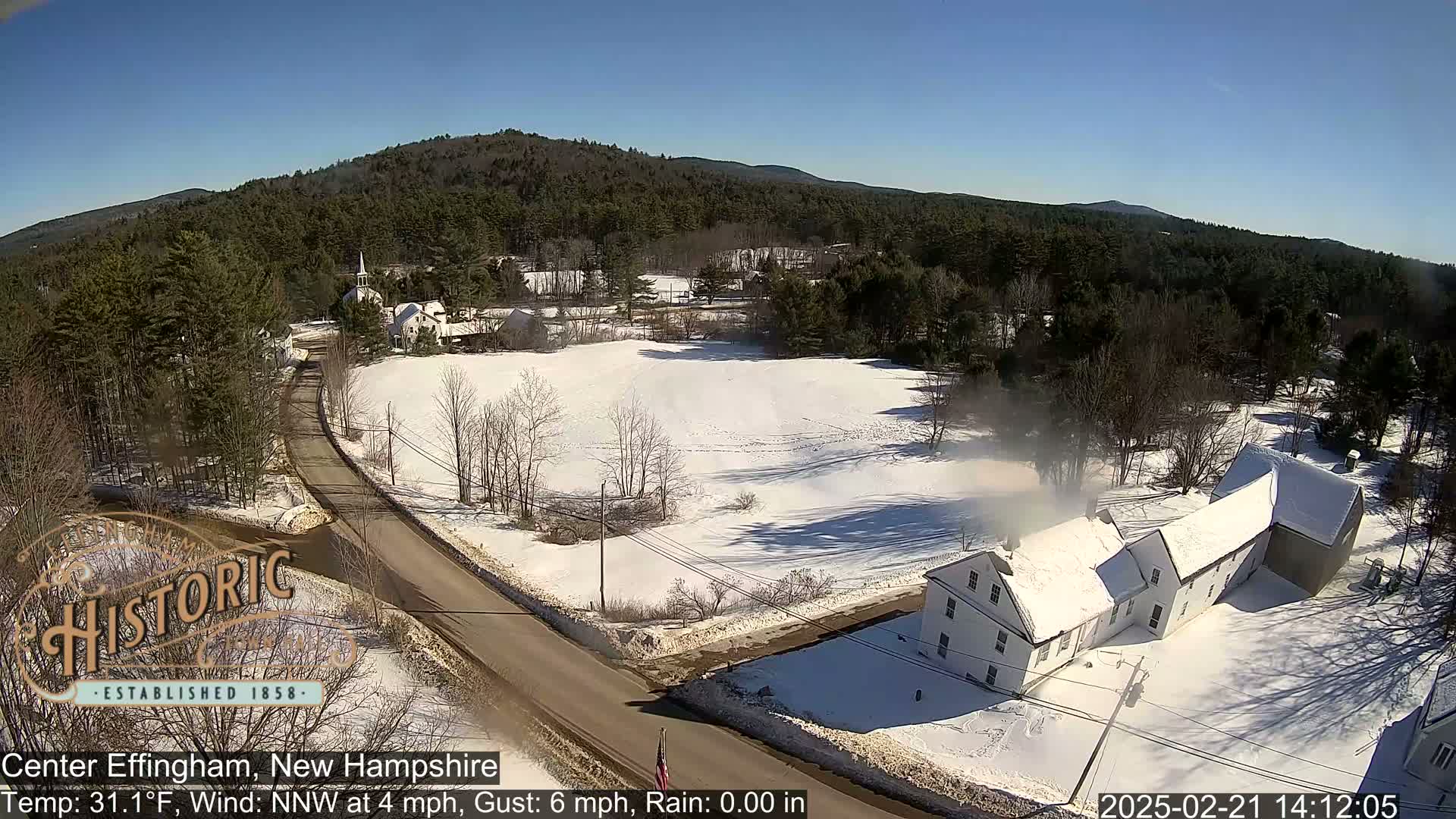A snow-covered road winds through a rural landscape of houses and trees under a clear blue sky.